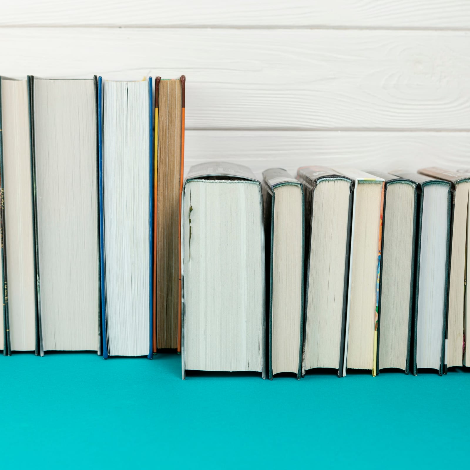 Fileira de livros de capa dura, cores variadas, dispostos lado a lado sobre superfície azul-esverdeada